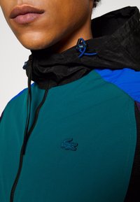 Teal and black lightweight jacket with a high collar, side zipper, and blue accents. Features an embroidered logo on the chest.