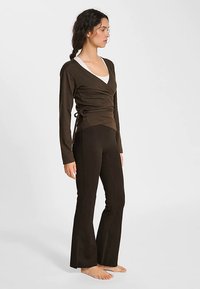 Brązowa bluzka z długimi rękawami i wiązaniami po bokach, zestawiona z ciemnobrązowymi rozszerzanymi spodniami do jogi. Tkanina wydaje się miękka i gładka.