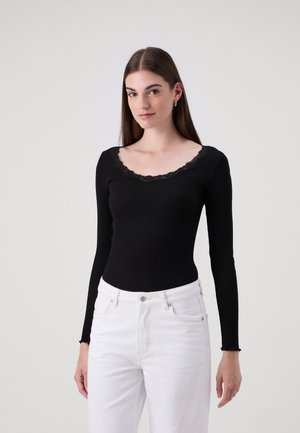 Mustat pitkät hihat bodysuit, jossa on ribbitekstuuri ja aallotettu, pitsireunainen kaula-aukko. Yhdistetty korkeavyötäröisiin valkoisiin farkkuihin.