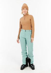 Bruine langemouwtop met rits, gecombineerd met mintgroene sneeuwbroeken met zwarte accenten en een hoge taille, gedragen met zwarte skischoenen.