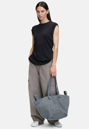 Graue Leinensh Tasche mit stabilen Griffen, präsentiert von einer Person in einem schwarzen Rippoberteil und grauen Cargo-Hosen, die ihr strukturiertes Design zur Schau stellt.