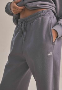Pantalones de chándal de algodón gris con cintura elástica, cordón y bolsillos laterales. Presenta el logo bordado 'self.' en blanco en la pierna izquierda.