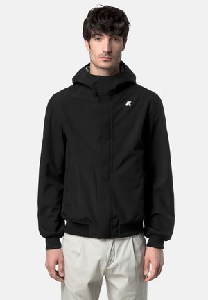 Uomo che indossa una giacca nera con cappuccio e logo sul petto e pantaloni chiari, in piedi davanti a uno sfondo neutro.