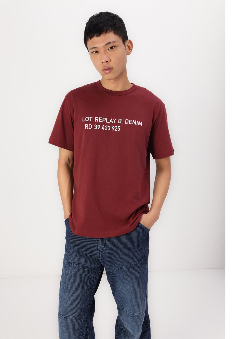 Bordeauxfarbenes T-Shirt aus Baumwolle mit kurzen Ärmeln, mit weißem Text "LOT REPLAY B. DENIM RD 39 423 925" quer über der Brust. Jeanskörper.