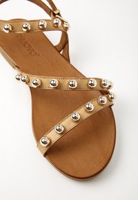 Sandale beige avec deux brides ornées de clous métalliques ronds, dotée d'une semelle plate et d'une bride de cheville ajustable.