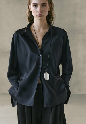 Kvinde iført en løs sort skjorte med knapper og sorte bukser, hænderne i lommerne, med en lang halskæde med en oval sølvvedhæng.