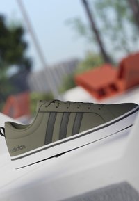 Zapatilla Adidas baja de color verde oliva con rayas negras y suela blanca, situada sobre una superficie blanca con un fondo exterior desenfocado.