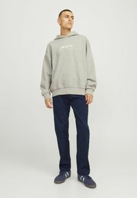 Mand stående iført en grå hættetrøje med "aprel"-logo, mørkeblå bukser og marineblå sneakers med hvide striber mod en ensfarvet baggrund.