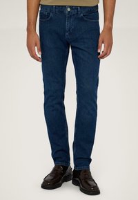 Jeans de mezclilla azul oscuro con un corte ajustado, que cuentan con cinco bolsillos y cierre de botón de metal. Se combinan con zapatos de cuero oscuros.