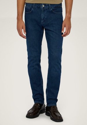Donkerblauwe denim jeans met een slim fit, voorzien van vijf zakken en een metalen knoopsluiting. Draagt samen met donkere leren schoenen.
