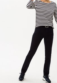 Persona che indossa una maglietta a righe bianche e nere a maniche lunghe, pantaloni neri con una cintura con fibbia argentata e sneakers nere, in piedi su uno sfondo bianco.