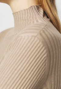 Sweter z prążkowanym kołnierzem w jasnym beżu, z wysokim kołnierzem i gładką teksturą, podkreślający dopasowaną sylwetkę z pionowymi paskami.