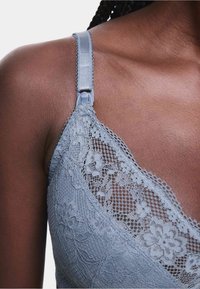 Bralette in pizzo grigio chiaro con motivi floreali, tessuto testurizzato, spalline regolabili e bordo ondulato lungo il collo.