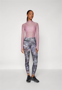 Hellviolettes Langarmoberteil mit Mesh-Details, kombiniert mit hoch taillierten Leggings mit dunklem Galaxie-Print, ergänzt durch schwarze Sportschuhe.