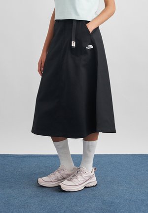 Inimene, kes kannab musta keskmise pikkusega seelikut The North Face logoga, valgeid sokke ja heleroosaid spordijalatseid, seisab sinisel vaibal.