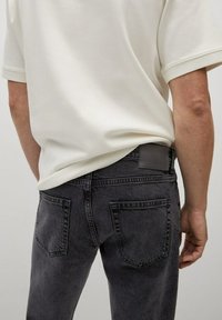Jeans in denim nero con una finitura sbiadita, dotati di due tasche posteriori e un'etichetta in pelle grigia alla vita. Indossati con una camicia chiara.