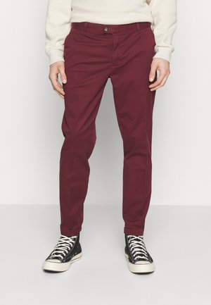 Man wearing dark red pants, a light beige long-sleeve shirt, and black high-top sneakers with white laces standing on a plain background.