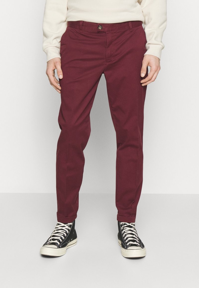 Homme portant un pantalon rouge foncé, une chemise beige clair à manches longues et des baskets montantes noires avec des lacets blancs, debout sur un fond uni.