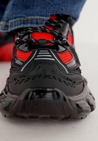 Schwarze und rote Sportschuhe aus glänzendem Synthetikmaterial, mit atmungsaktiven Mesh-Panels, dicken Sohlen und einem detaillierten Schnürdesign.