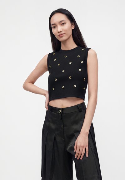 Femme portant un crop top noir sans manches avec des détails œillets dorés et un pantalon noir taille haute à jambes larges, debout sur un fond blanc.