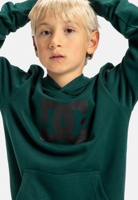 Zelená mikina s kapucí, na přední straně s velkým černým logem DC. Vyrobena z měkké tkaniny s klokankou.