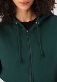 Sudadera verde con cremallera y capucha ajustable, que presenta detalles en metal plateado y una textura suave. La vista en primer plano destaca la cremallera y los detalles de la tela.