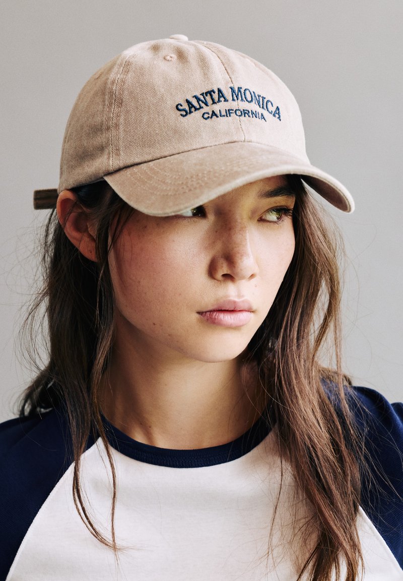 Jeune femme aux longs cheveux bruns portant une casquette beige Santa Monica Californie et une chemise bleu marine et blanche, regardant sur le côté sur un fond uni.