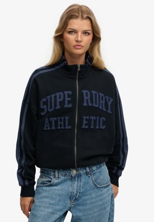 Ung kvinde med langt blondt hår iført en sort lynlåstrøje med teksten "Superdry Athletic" og blå stribedetaljer, kombineret med lyseblå jeans.