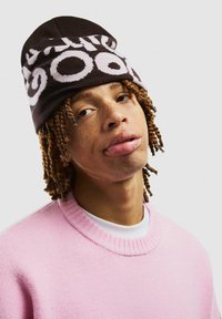 Ruskea neulepipo, jossa on valkoista tekstiä, yhdistettynä vaaleanpunaiseen neulepaitaan. Rakenne on teksturoitu, muoto on pyöreä ja ulkoasu kerroksellinen.