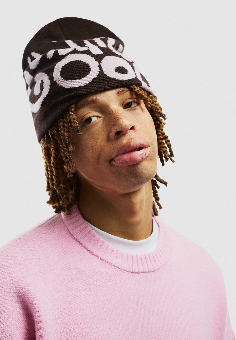 Ruskea neulepipo, jossa on valkoista tekstiä, yhdistettynä vaaleanpunaiseen neulepaitaan. Rakenne on teksturoitu, muoto on pyöreä ja ulkoasu kerroksellinen.