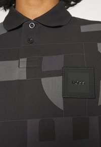 Polo noir avec un motif géométrique en nuances de gris. Tissu texturé avec une petite étiquette noire portant la mention "BOSS" sur la poitrine.