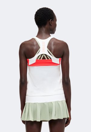Mujer con una camiseta deportiva sin mangas blanca y roja con espalda cruzada y "ENDLESS" en la espalda, y una falda plisada verde claro, vista desde atrás.