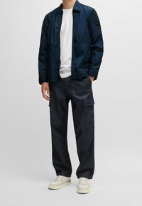 Marinblå lättviktsjacka med dragkedja, som kombineras med mörka denim cargo-byxor med sidofickor och krämfärgade sneakers.