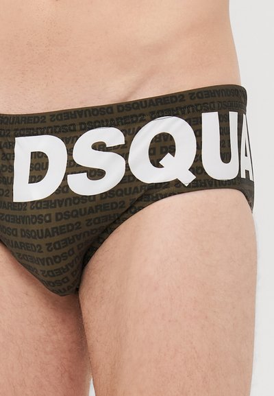 Zoom na osebi, ki nosi temno zelene kopalne hlačke z ponavljajočim se vzorcem besedila "DSQUARED2" in velikimi belimi črkami "DSQUA" na sprednji strani.