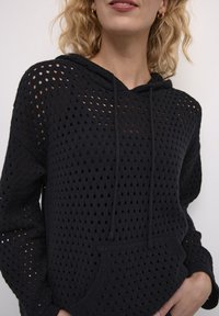 Melna adīta kapuce ar pārklājuma rakstu, kurai ir kangoru kabata un saites kapuce. Tekstūra ir brīva un elpojoša.