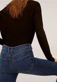 Camicia scura a maniche lunghe abbinata a jeans in denim blu aderenti con due tasche posteriori e vita a media altezza. Il denim ha una leggera elasticità.