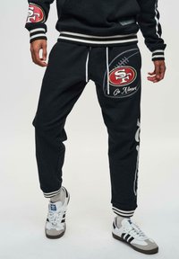 Zwarte joggers met een voetbaldessin, logo-opdruk en witte koordjes. Heeft gestreepte manchetten en een loose fit, gecombineerd met witte sneakers.