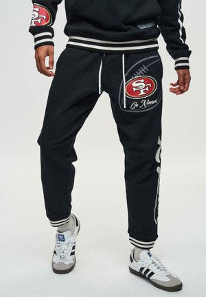 Svarta joggers med fotbolls grafiskt tryck, logotypapplikation och vita snörningar. Har randiga muddarna och en avslappnad passform, i kombination med vita sneakers.