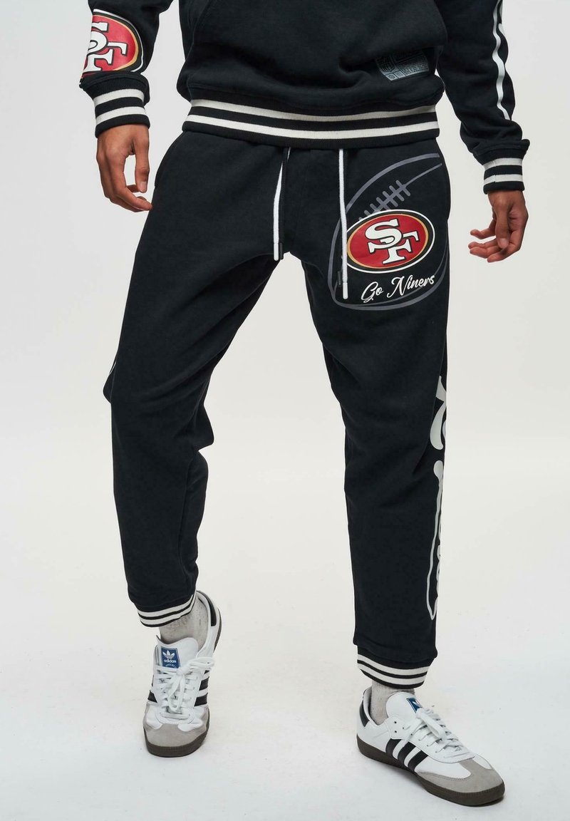 Zwarte joggers met een voetbaldessin, logo-opdruk en witte koordjes. Heeft gestreepte manchetten en een loose fit, gecombineerd met witte sneakers.