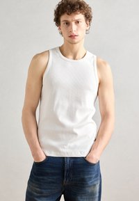 Débardeur blanc à côtes avec un col rond, design ajusté et texture lisse, associé à un jean bleu foncé. Le mannequin se tient avec les mains dans les poches.