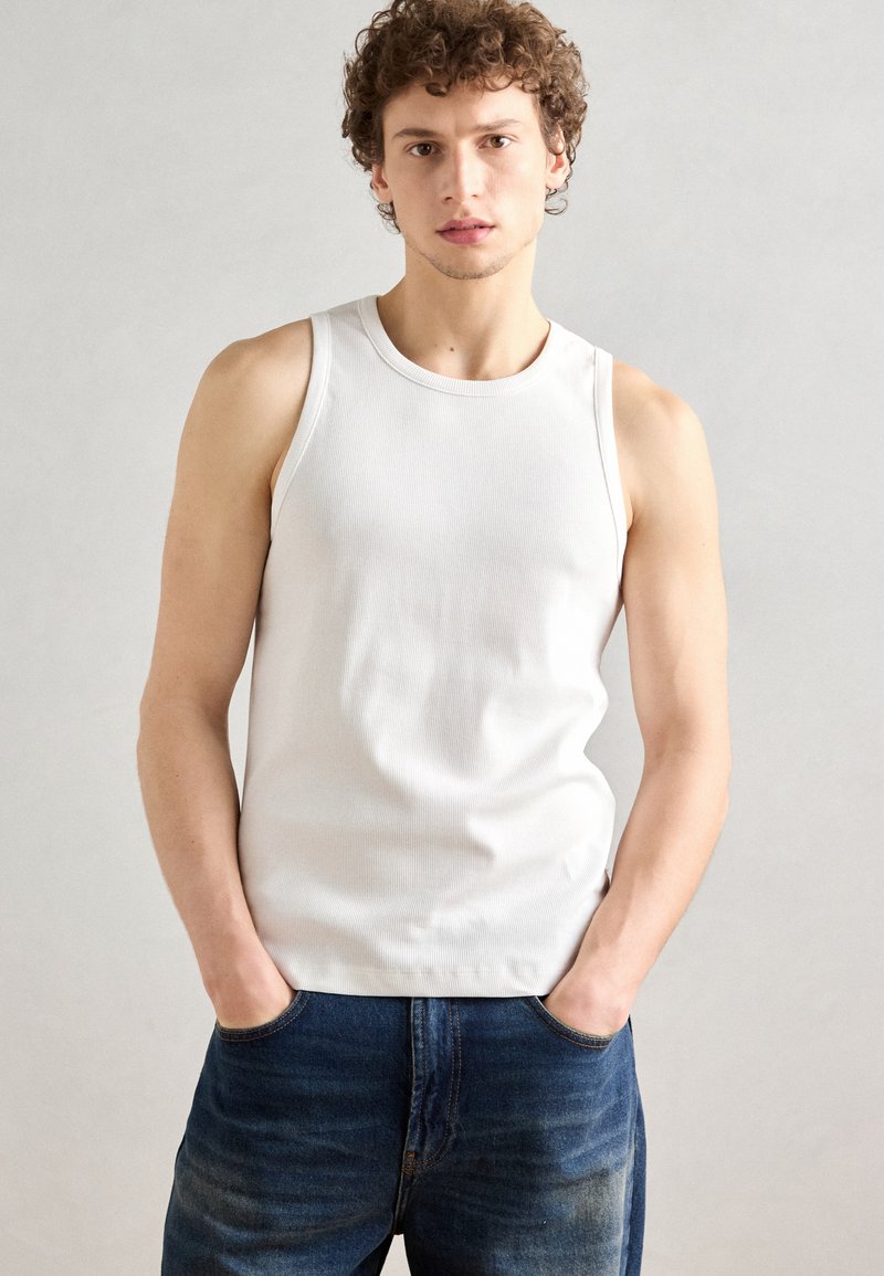 Débardeur blanc à côtes avec un col rond, design ajusté et texture lisse, associé à un jean bleu foncé. Le mannequin se tient avec les mains dans les poches.