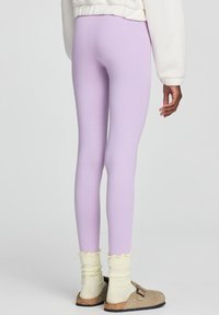 Leggings lilla realizzati in tessuto elastico, con un design aderente. Accompagnati da calzini giallo chiaro con bordo ondulato e scarpe beige senza lacci.