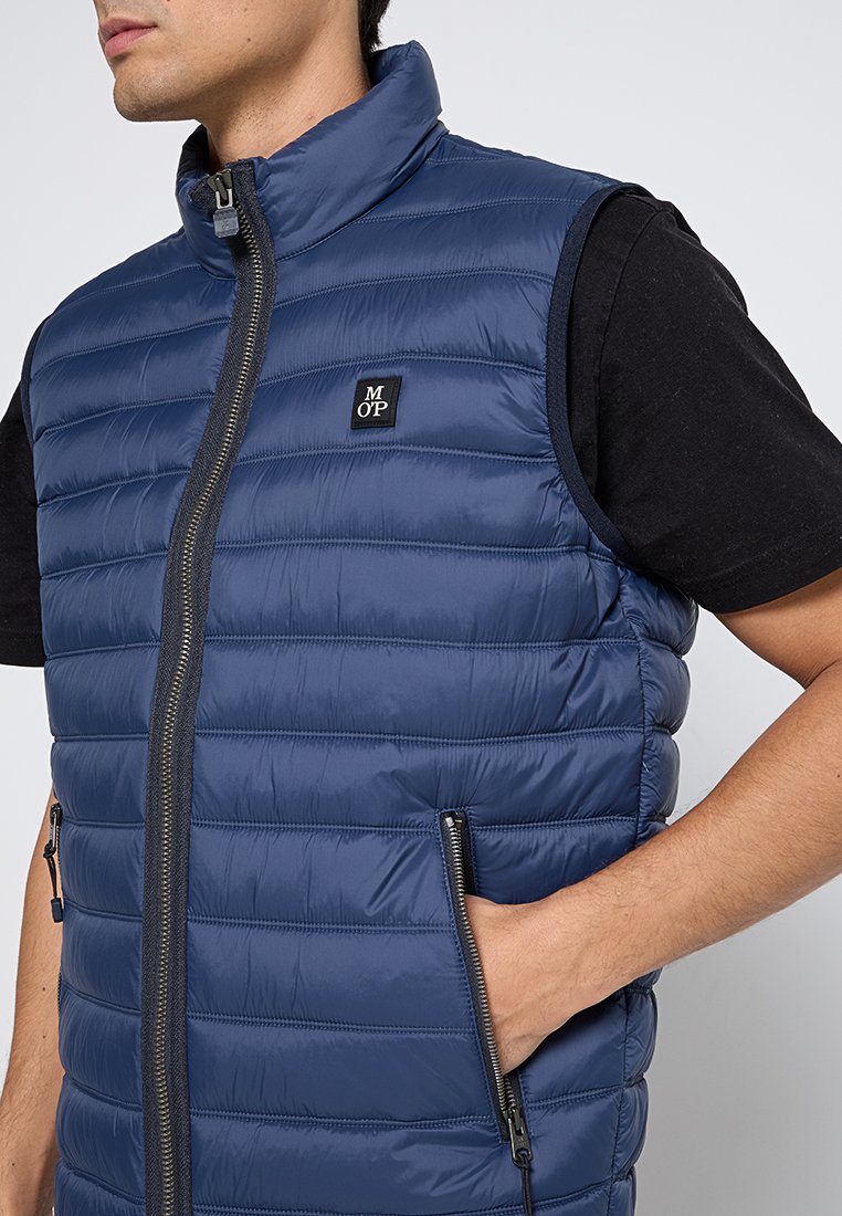 Granatowa kamizelka puchowa z pikowanym wzorem, z wysokim kołnierzem i zapięciem na zamek z przodu. Posiada boczne kieszenie na zamek oraz małą naszywkę z logo.