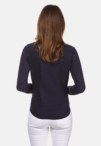 Camicia a maniche lunghe blu navy con una texture liscia, caratterizzata da un orlo curvo e polsini con bottone, indossata con pantaloni bianchi. Vista posteriore.
