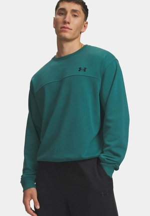 Junger Mann trägt einen türkisfarbenen Under Armour Pullover und schwarze Under Armour Hose, steht mit einer Hand in der Tasche vor weißem Hintergrund.