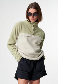 Fleece-Pullover in Hellgrün und Creme mit einem halben Reißverschluss, geknöpftem Kragen und Fronttasche. Kombiniert mit schwarzen Shorts im entspannten Schnitt.
