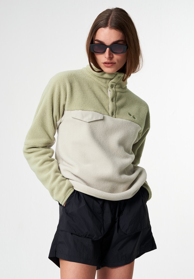 Fleece-Pullover in Hellgrün und Creme mit einem halben Reißverschluss, geknöpftem Kragen und Fronttasche. Kombiniert mit schwarzen Shorts im entspannten Schnitt.