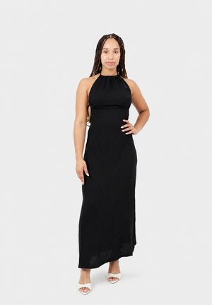 Femme aux longues tresses se tenant face à l'avant, vêtue d'une robe longue noire sans manches à col halter et de sandales blanches à bout ouvert, sur un fond uni.
