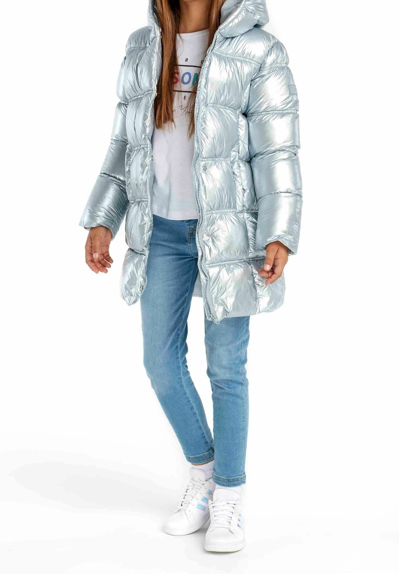 Personne portant une doudoune argentée brillante à capuche, un t-shirt blanc à motif graphique, un jean bleu et des baskets blanches avec des rayures bleu clair.