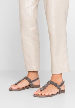 Voeten met beige cropped broek en donkergrijze platte sandalen met kralen en golvende bandjes en enkelgespen op een lichtgrijze vloer.